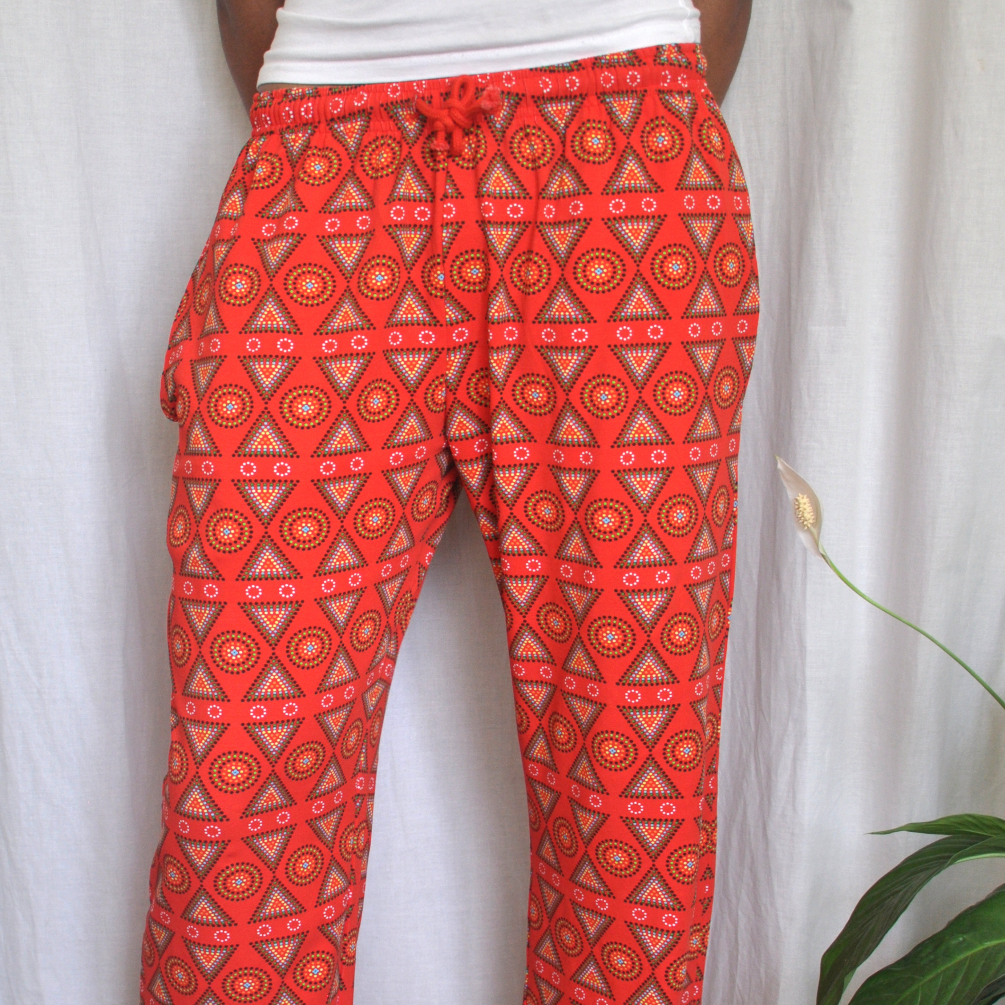 A person wearing vibrant, red, patterned pyjama pants with geometric designs stands in front of a white background. A small part of a green plant with a single white bloom is visible on the right side of the image.