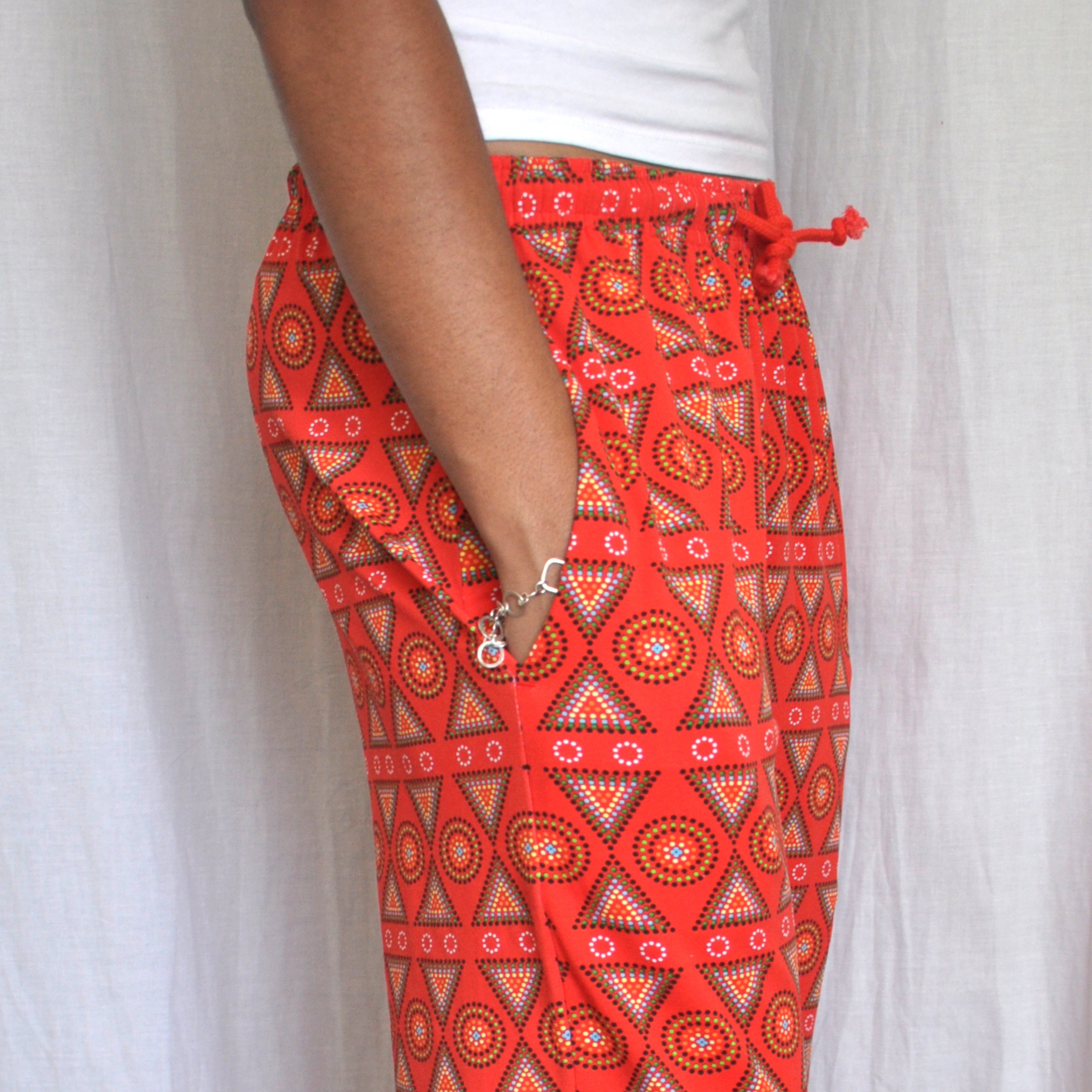 A person is standing in front of a white curtain, wearing a white top and red pyjama pants with a colorful geometric pattern. Their hand is casually placed in the pocket of their pyjama pants, revealing a silver bracelet on their wrist.