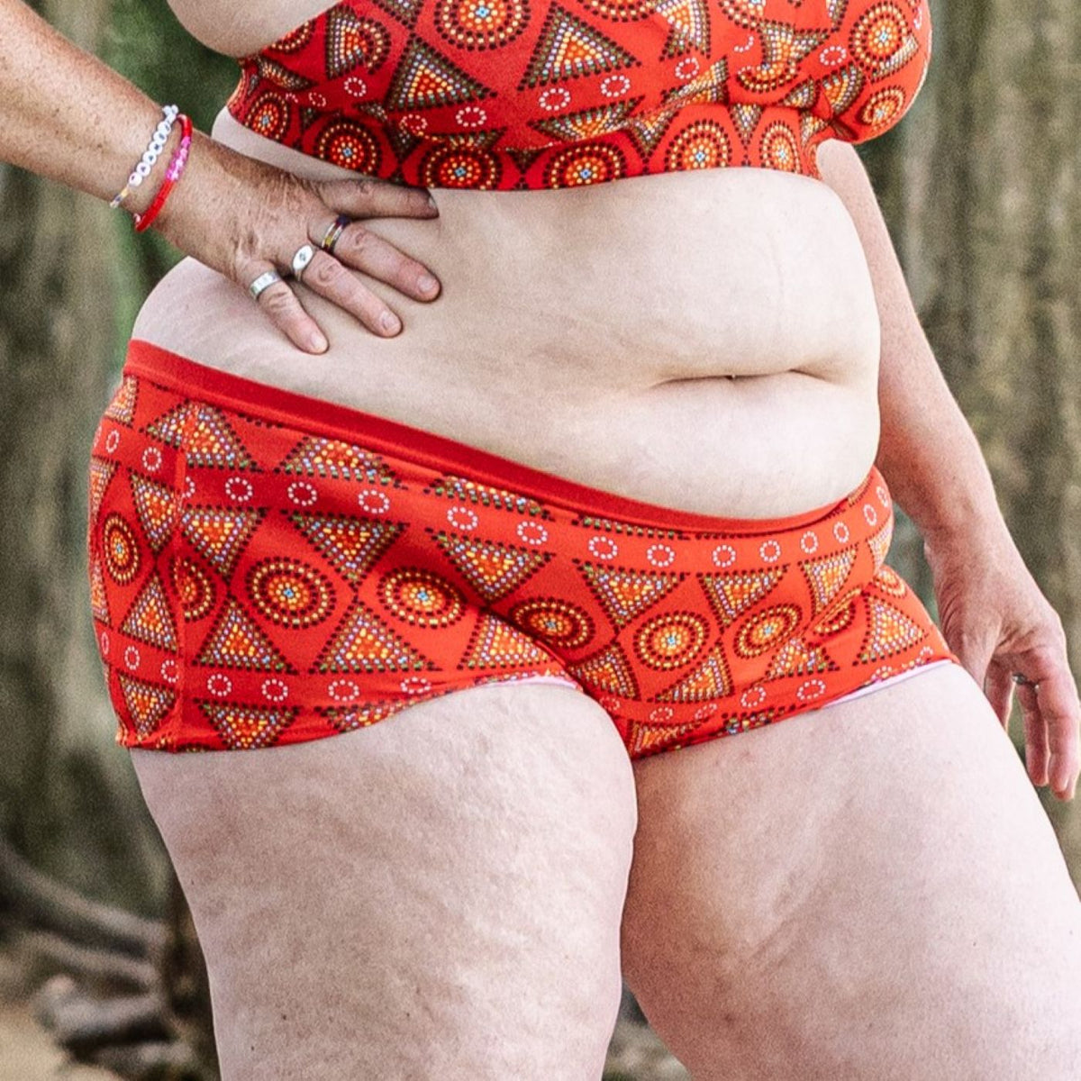 A photo of a model wearing the YOU Underwear boyshorts in red mara. The photo is zoomed in so we can see from the model's stomach to their mid-thigh. They are facing the camera with their right hand on their hip. 