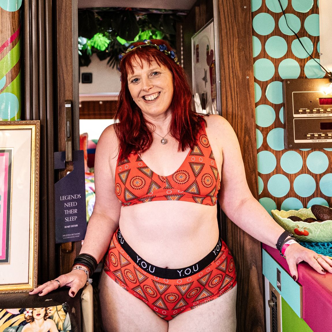 A photo of a model wearing the YOU Underwear bralette and branded boyshorts in the red mara print. She is standing in a doorway, with her hands of two table surfaces either side of her. She is standing facing the camera and smiling. She is wearing a flower crown on her head and there is a wall with big blue polka dots on it to her right.