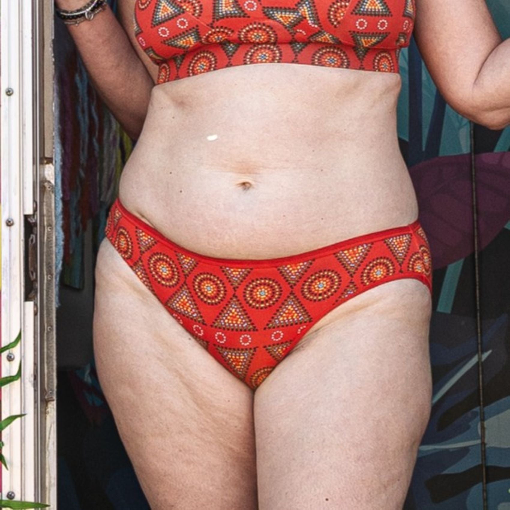 A photo of a model wearing the YOU Underwear low rise bikini knickers in the vibrant red mara print. The photo is zoomed in so we can see from the model's upper stomach to their mid-thigh. They appear to be stood in a doorway.