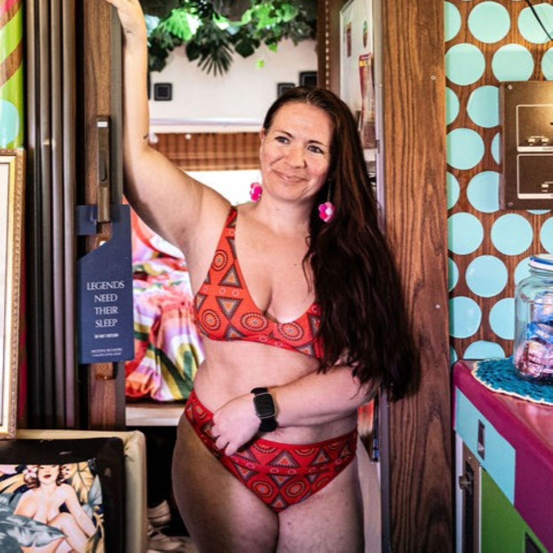 A photo of a model wearing the YOU Underwear red mara bralette and mid-rise bikini knickers. She is standing in a doorway, smiling and looking out to the left. There is a bright blue polka dot wall on the left and green foliage coming from the ceiling in the background.