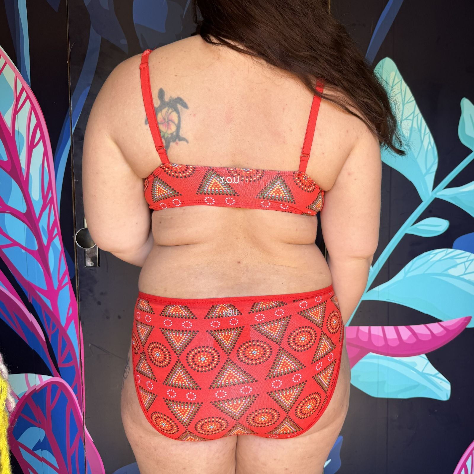 A photo of a model wearing the YOU Underwear red mara bralette and matching mid-rise knickers. The photo is zoomed in so we can see from the model's shoulders to their lower thigh. They are facing away from the camera. There is a black wall with bright blue and pink leaves on as the background.