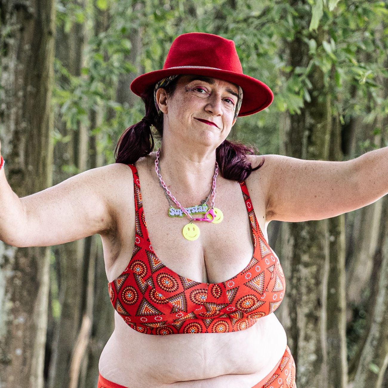 A photo of a model wearing the YOU Underwear non wired bra in the red mara print. She is always wearing a chunky, charm necklace and a red hat. She is facing the camera, with her arms out in front of her. There are lots of trees in the background.