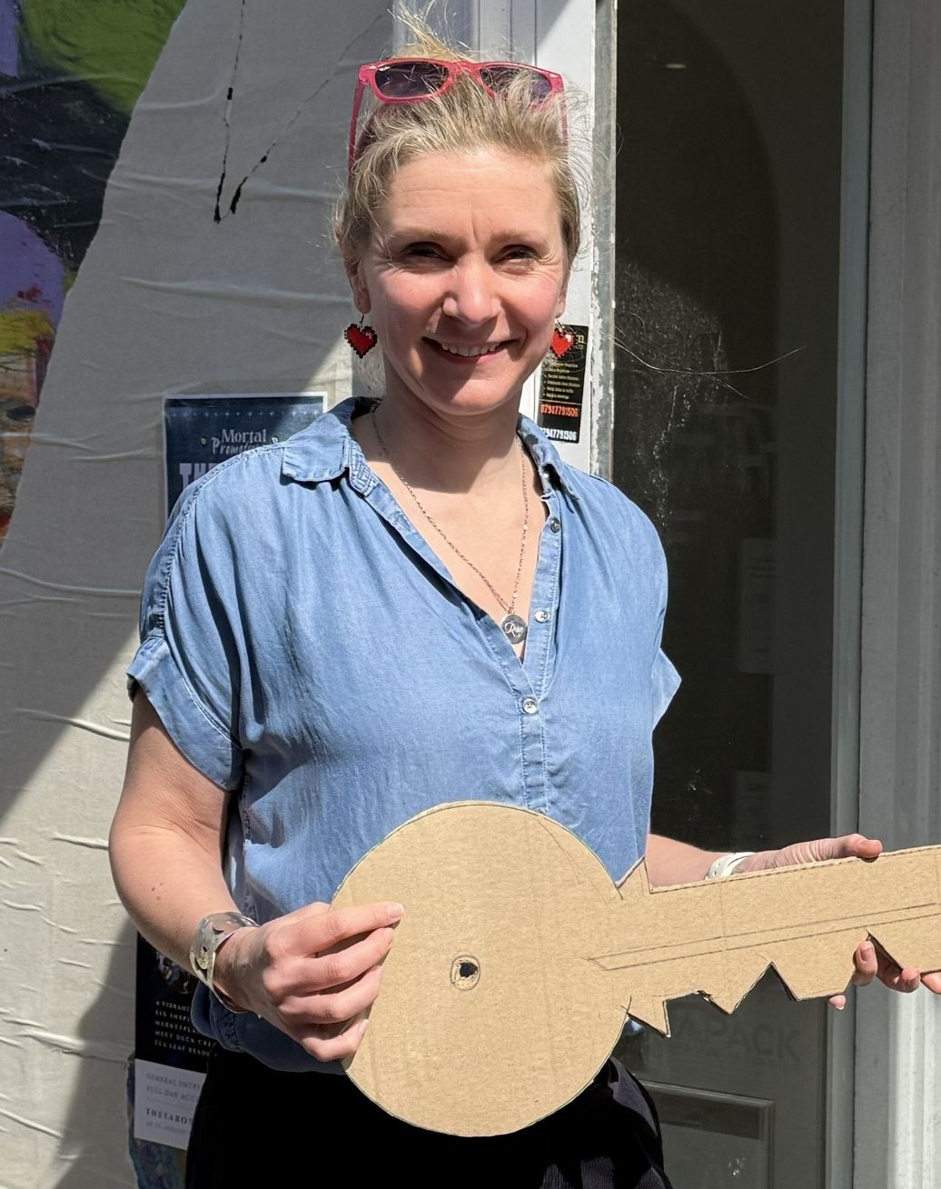 Photo shows Sarah, a lady in a blue shirt, standing outside a white building with a large cardboard key. 