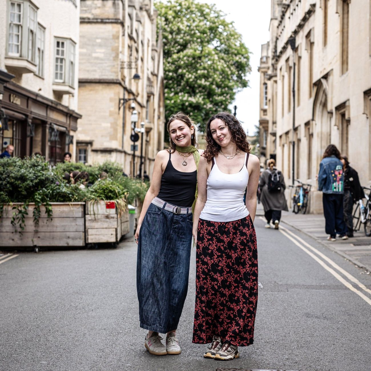 A picture of two women standing on a city street, outside. One is wearing the YOU Underwear black camisole with jeans and the other is wearing the white camisole with a black and red flowery skirt. They are facing the camera, smiling. There are big stone buildings behind them and some green plants on their left.