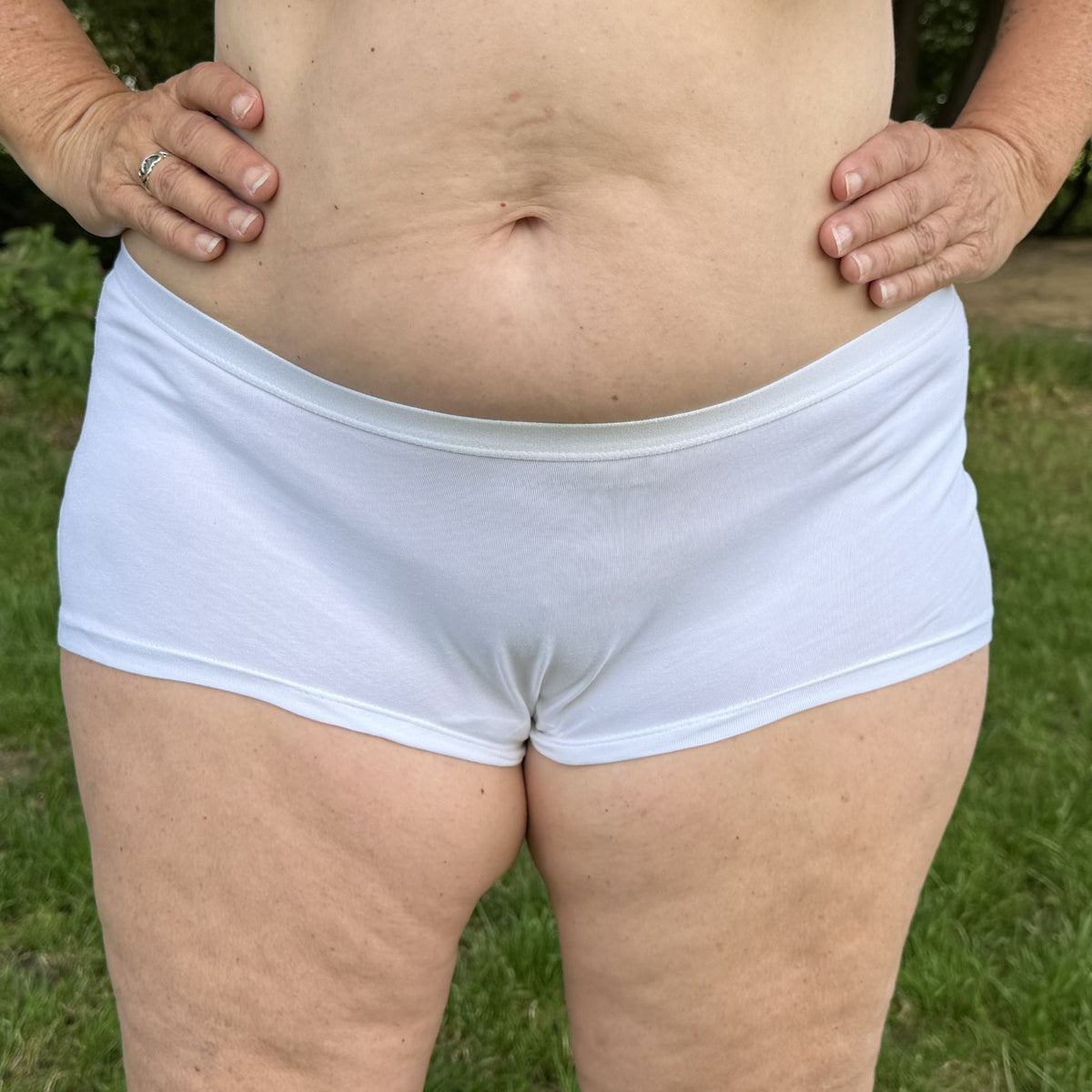 A photo of a model wearing the YOU Underwear boyshorts in white. The photo is zoomed in so we can see from the model's mid-stomach to their upper-thigh. They are facing the camera and have their hands on their hips. In the background there is green grass.