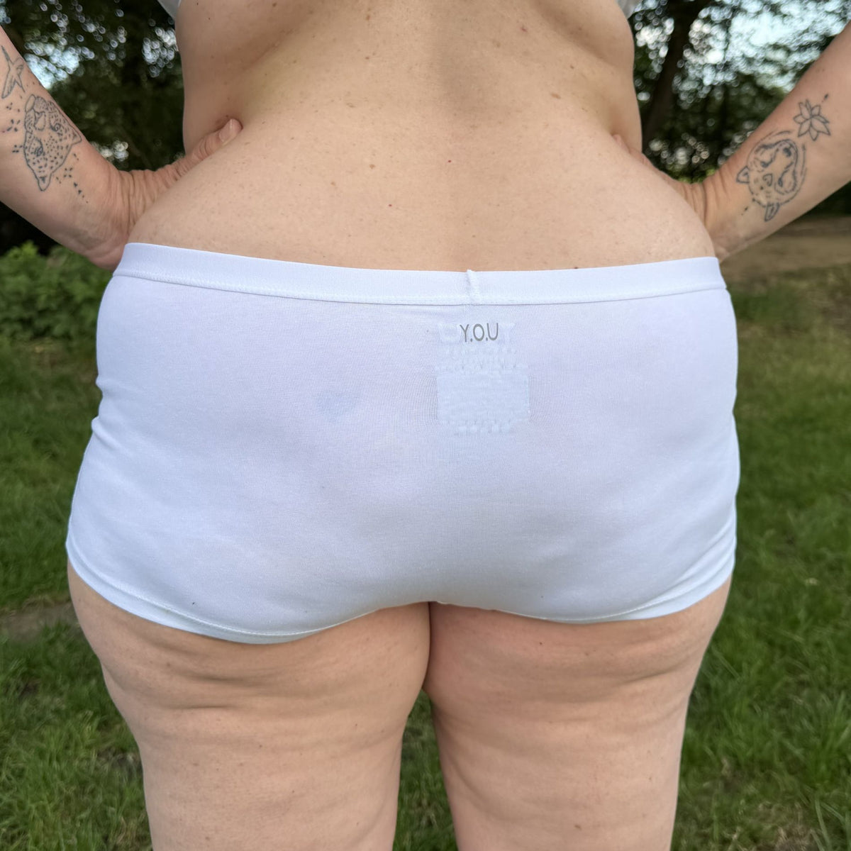 A photo of a model wearing the YOU Underwear boyshorts in white. The photo is zoomed in so we can see from the model's back to their upper-thigh. They are facing away from the camera and have their hands on their hips. In the background there is green grass.