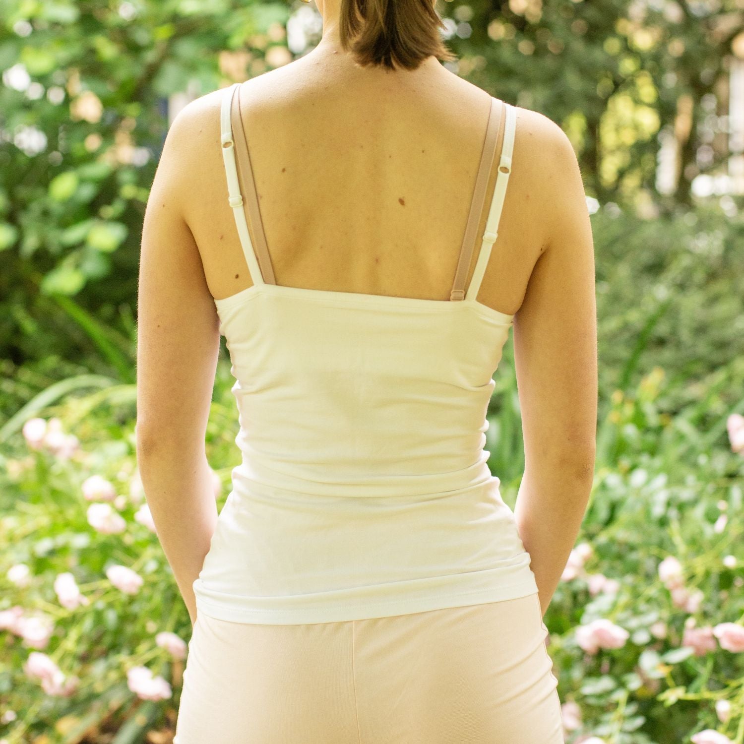 Product focused image of a model wearing the YOU Underwear Organic Cotton White Camisole. The model faces away from the camera, with her arms by her side. She stands in front of a bush with pink flowers. The image is cropped from the chin to the hips.