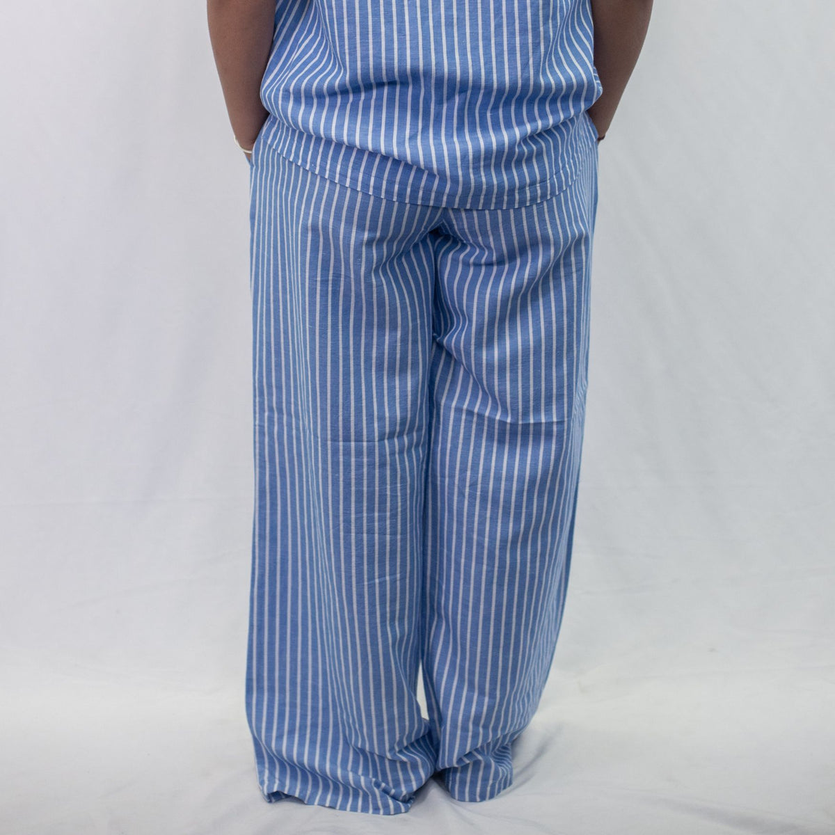 A photo of a model wearing the YOU Underwear woven pyjama bottoms in the classic blue and white stripe. The photo is zoomed in so we can see from the model's stomach to the floor. They are standing facing away from the camera, with their hands in their pockets, and there is a white background in front of them.