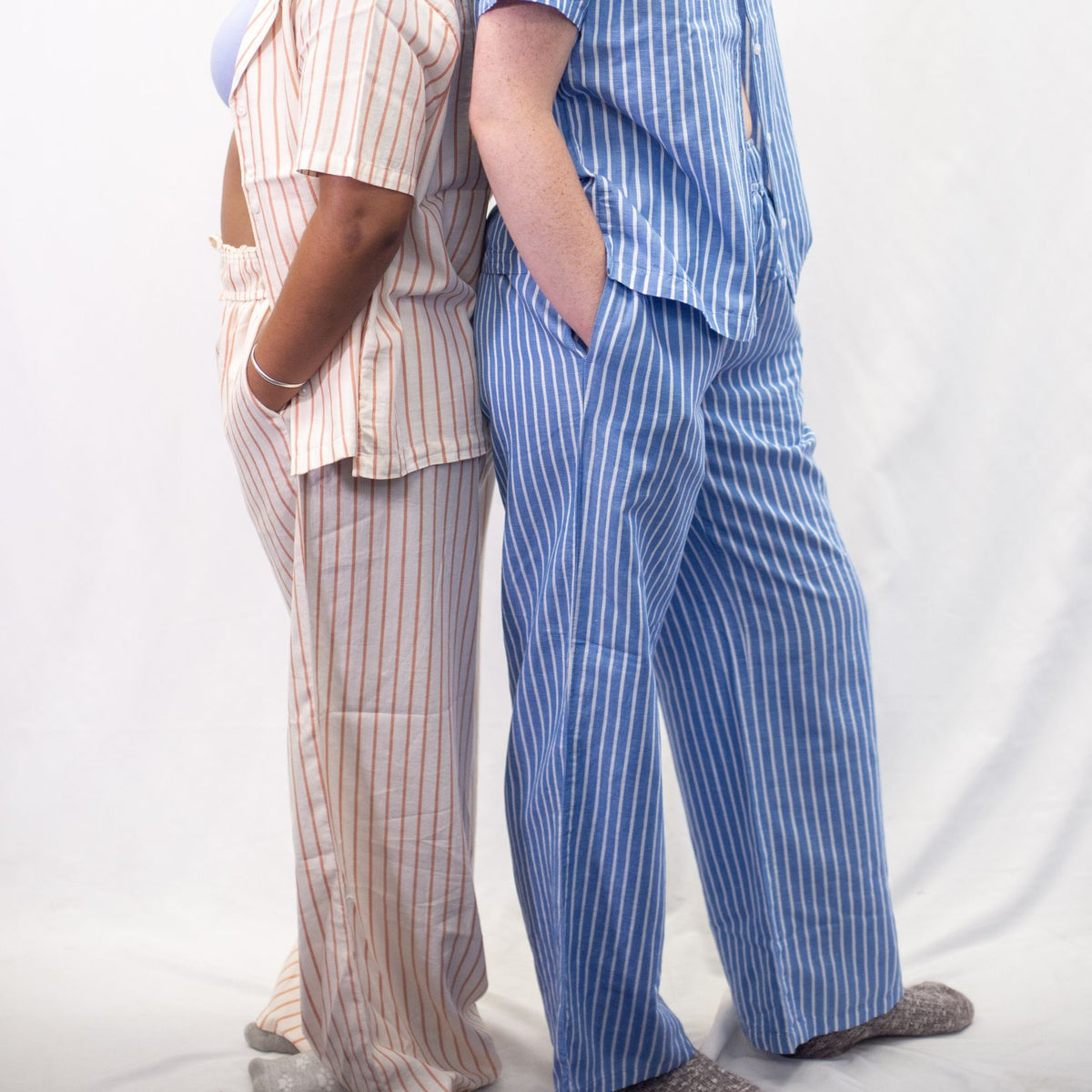 A photo of two models wearing the YOU Underwear woven striped pyjamas. They are standing back to back, side on to the camera with their hands in their pockets. The photo is zoomed in so we can see from the model's chest to the floor. The model on the left is wearing the cream and coral stripes and the model on the right is wearing the blue and white stripes.