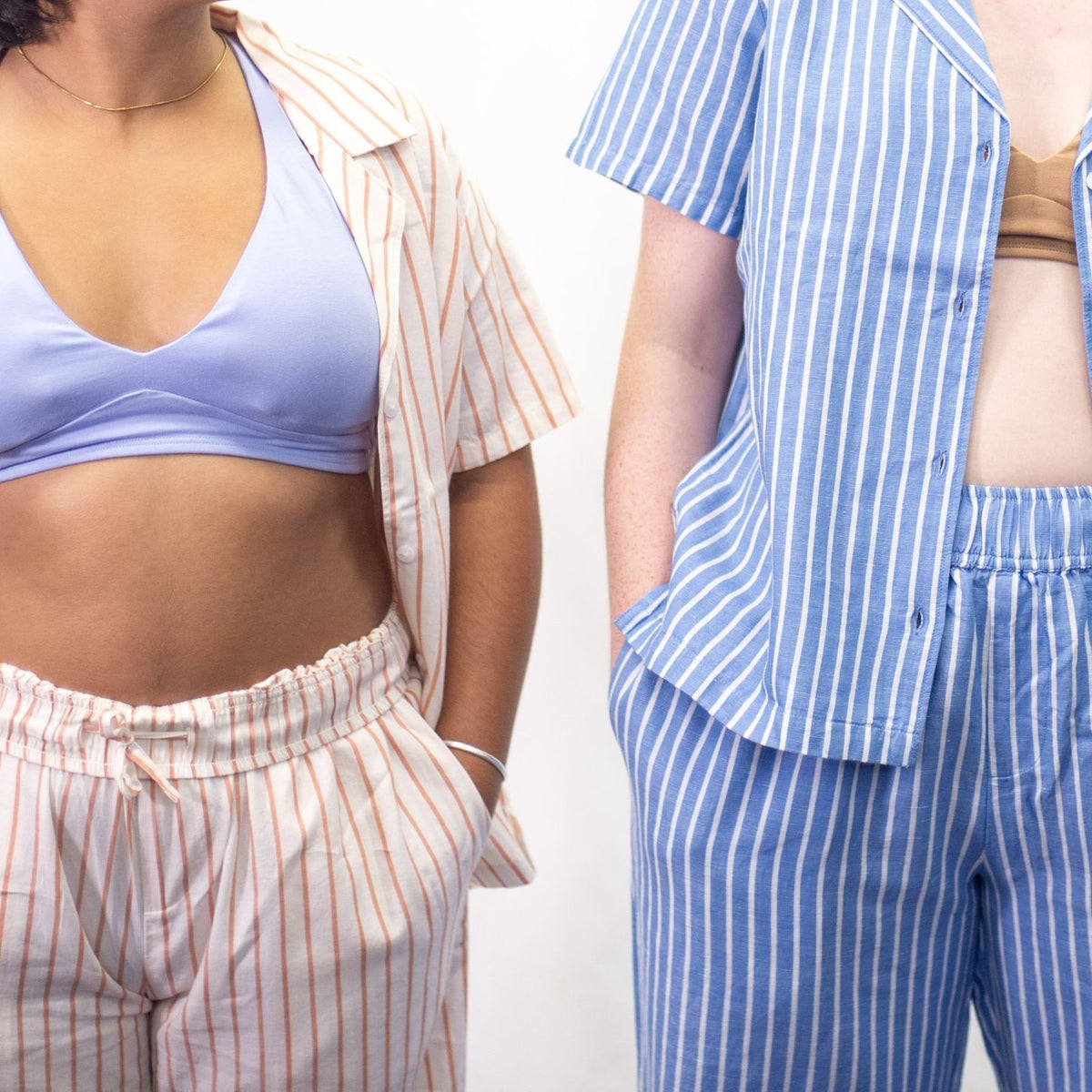 A photo of two models wearing the YOU Underwear woven striped pyjamas. The photo is zoomed in so we can only see from the model's shoulders to their thighs. The model on the left is wearing the soft cream and coral woven pyjamas, the shirt open to show a light blue YOU Underwear bralette. The model on the right is wearing the classic blue and white striped pyjamas, with the shirt unbuttoned to show the almond YOU Underwear bralette. 