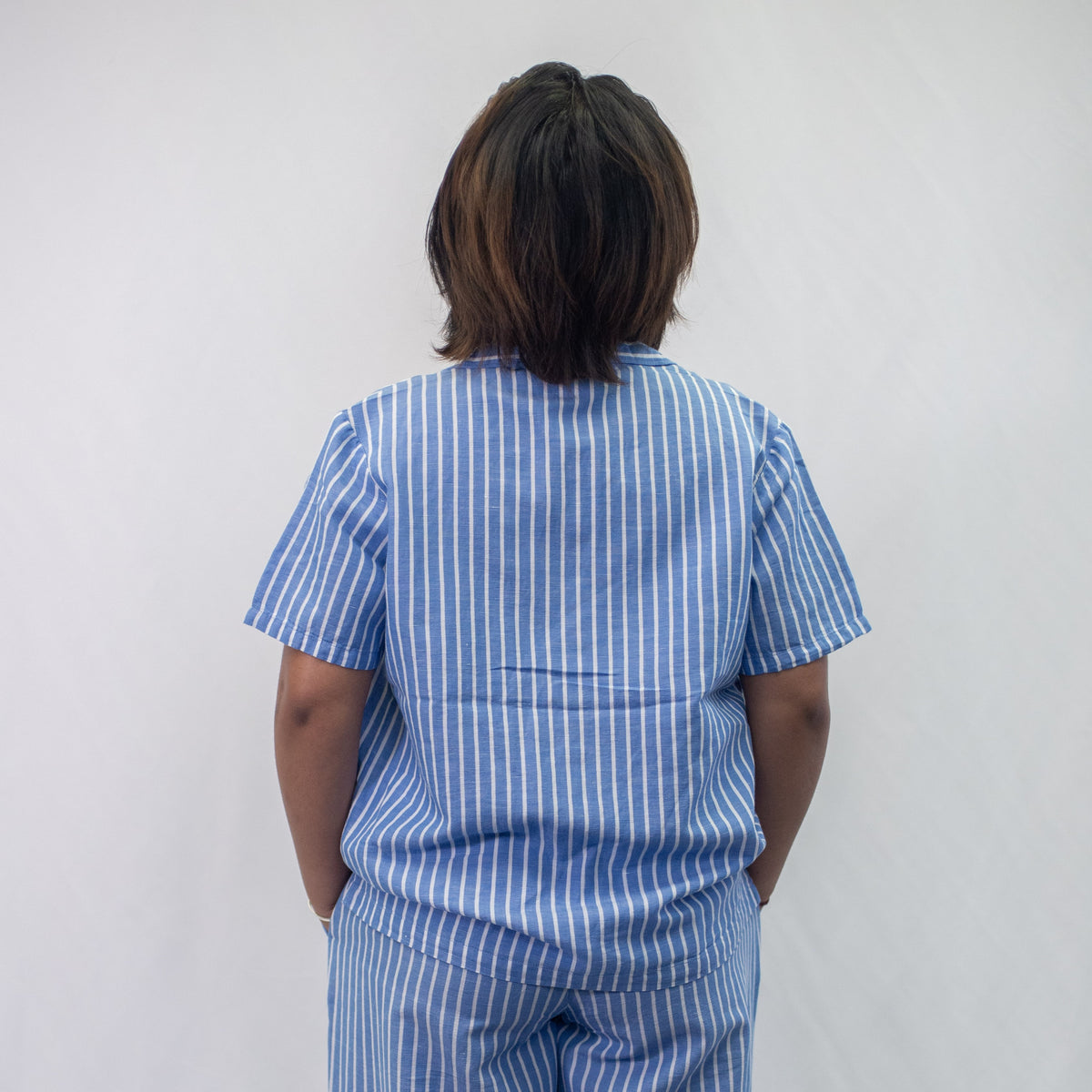A photo of a model wearing the YOU Underwear woven pyjama shirt in the classic blue and white stripe. She is standing facing away from the camera. There is a white background and she appears to have her hands in her pockets.
