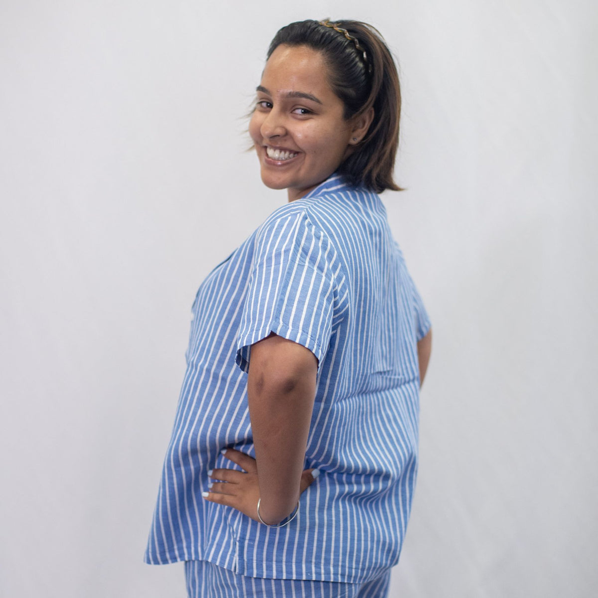 A photo of a model wearing the YOU Underwear woven pyjama shirt in the classic blue and white stripe. She is standing side on to the camera but looking into the lens and smiling with her hands on her hips. There is a white background behind her.