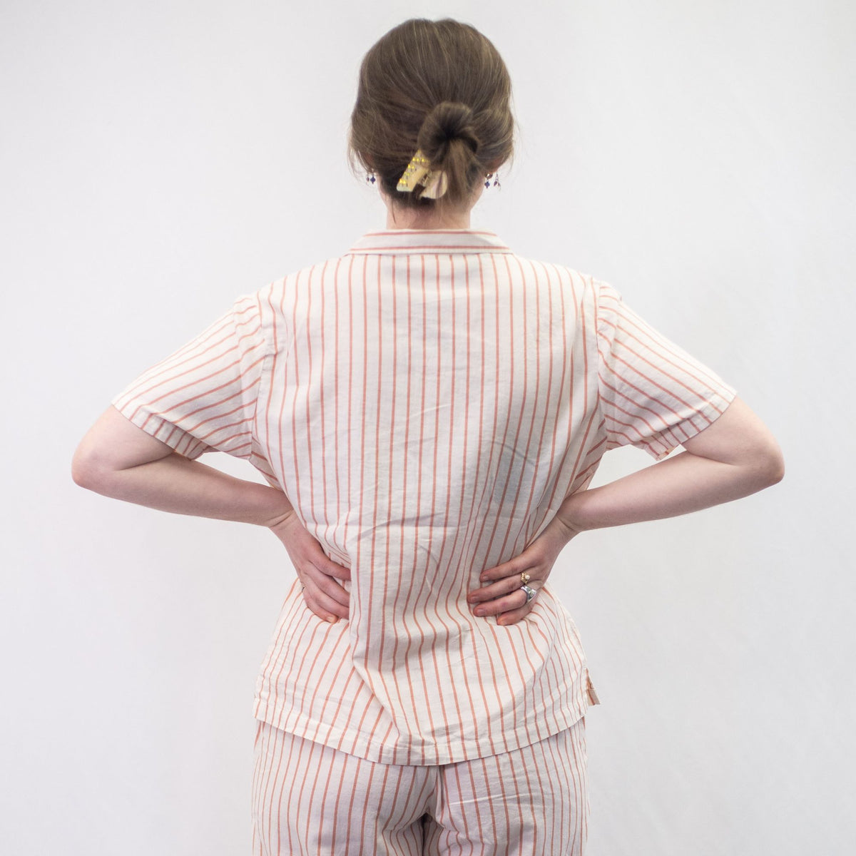 A photo of a model wearing the YOU Underwear woven pyjama shirt in the soft cream and coral stripe. She is standing facing away from the camera with both hands on her hips. There is a white background. 