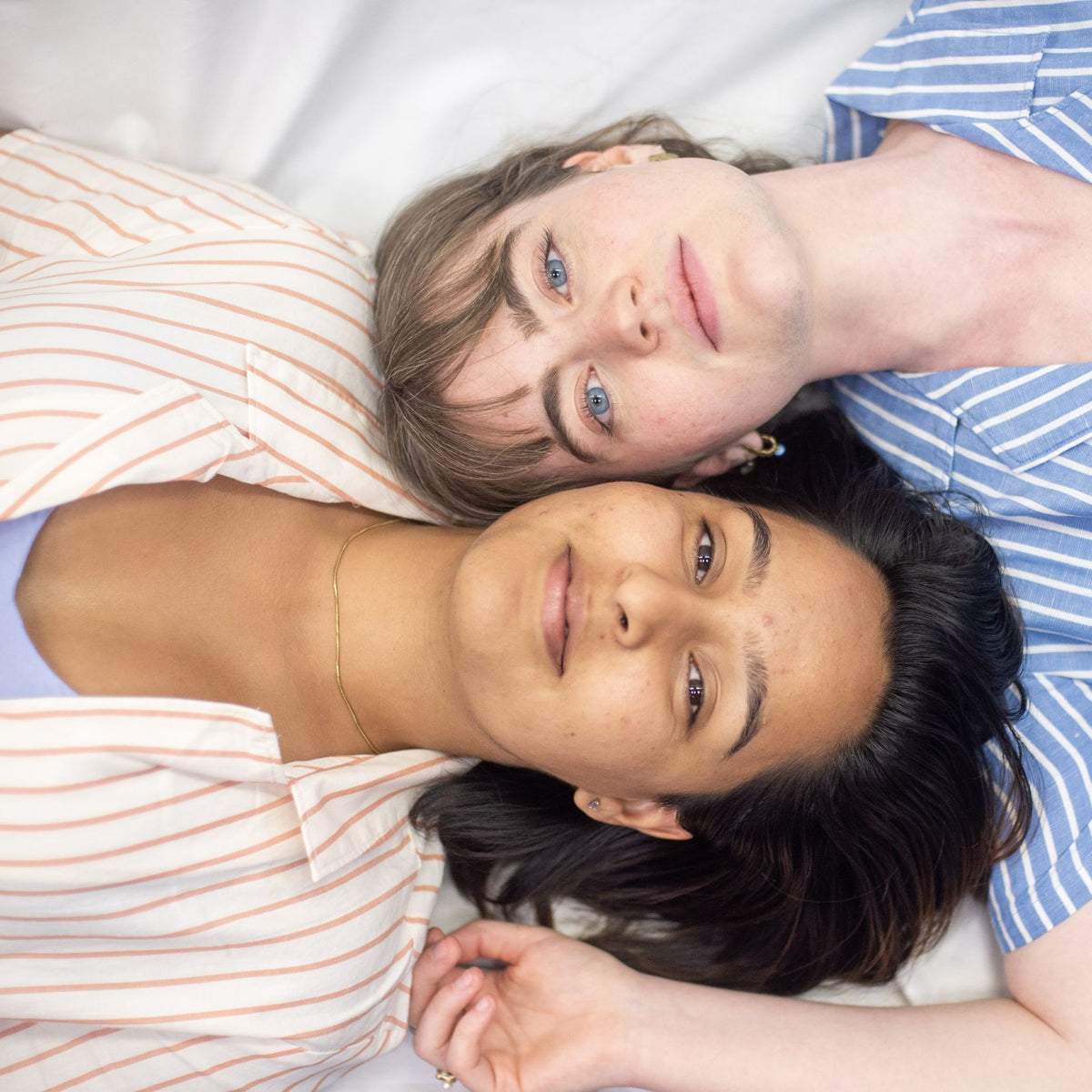 A birds-eye photo of two models laying down next to each other. The model on the left is wearing the shirt is soft cream and coral stripes and the model on the right is wearing the classic blue and white stripes. They are softly smiling and looking into the camera lens.