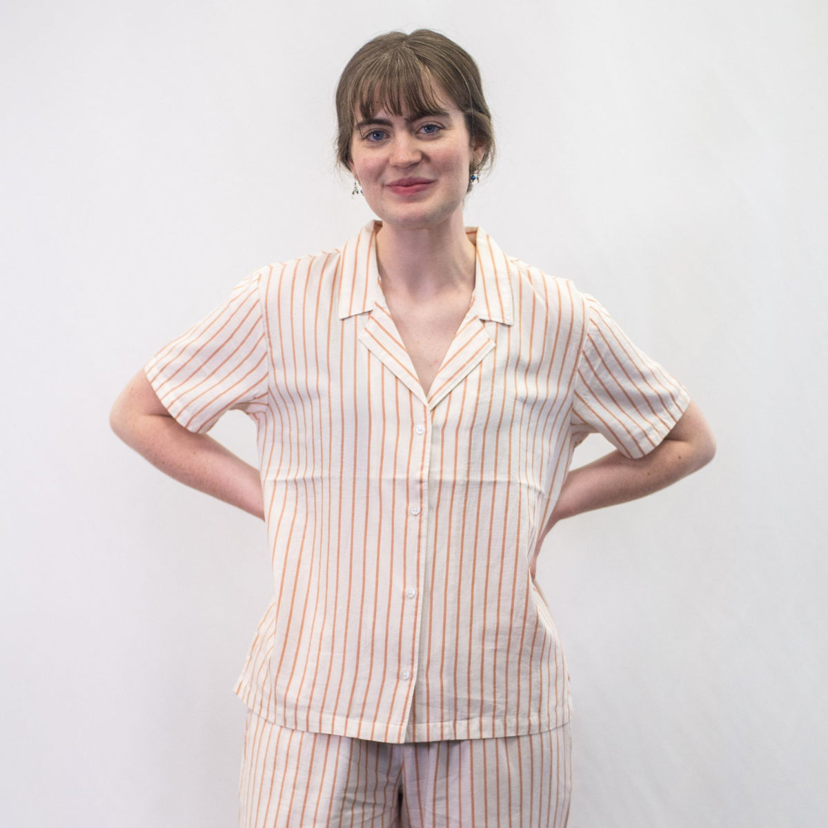 A photo of a model wearing the YOU Underwear woven pyjama shirt in the soft cream and coral stripe. She is standing face on to the camera with both hands on her hips. There is a white background. 