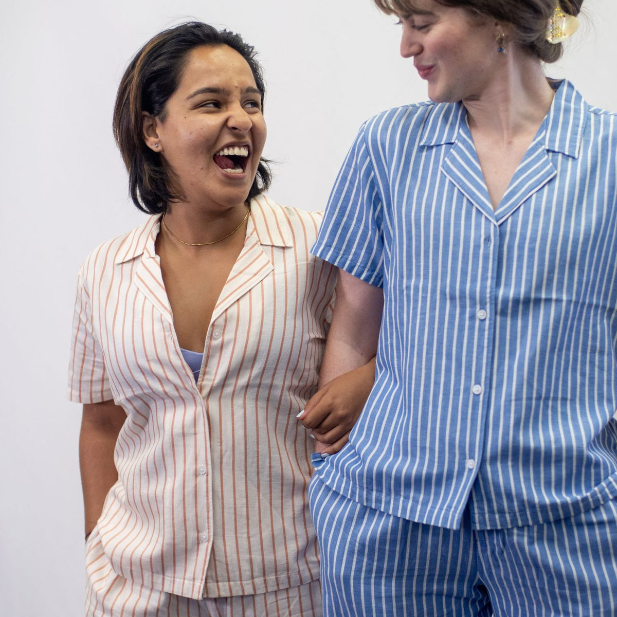 A photo of two model wearing the YOU Underwear woven pyjama shirts. The woman central in the photo is wearing the shirt in soft cream and coral stripes. She is facing the camera and linking arms with a model wearing the pyjama shirt in the classic blue and white stripes. 