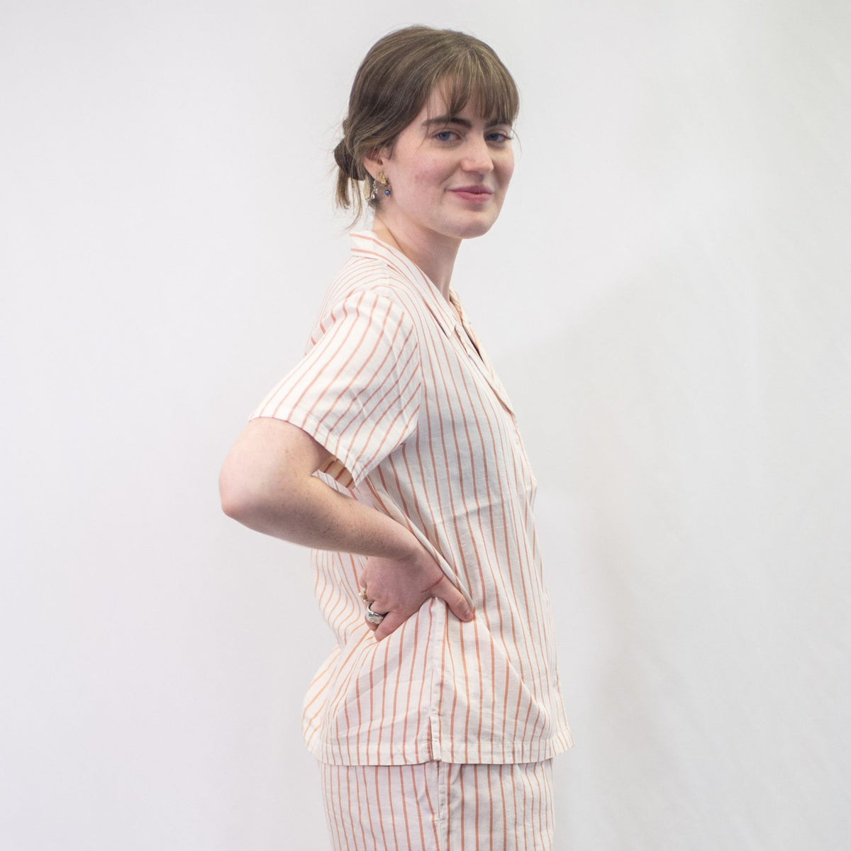 A photo of a model wearing the YOU Underwear woven pyjama shirt in the soft cream and coral stripe. She is standing side onto the camera with both hands on her hips. There is a white background. 