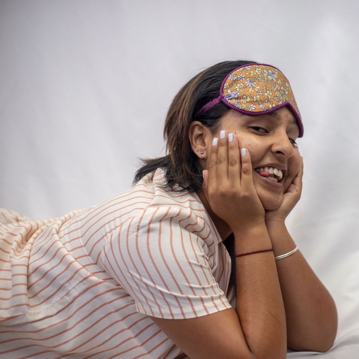A photo of a model wearing the YOU Underwear woven pyjama shirt in soft cream and coral stripes. She is laying n her front, propping her head up on her hands and cheekily smiling at the camera with her tongue out. She is wearing an orange and purple flowery sleep mask on her forehead. There is a white background.