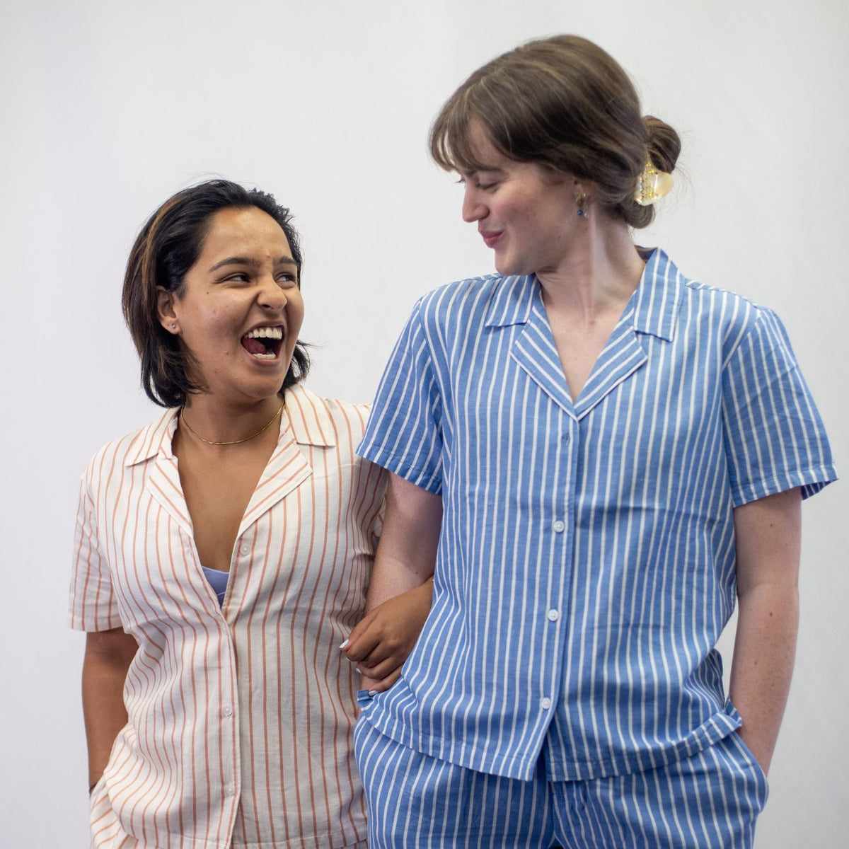 A photo of two women who are wearing the YOU Underwear woven striped pyjama shirts and matching trousers. They are standing facing the camera, linking arms and looking at each other in laughter. The woman on the left is wearing the soft cream and coral striped pyjamas and the woman on the right is wearing the classic blue and white stripes. They have their other hand in their pockets and there is white background.