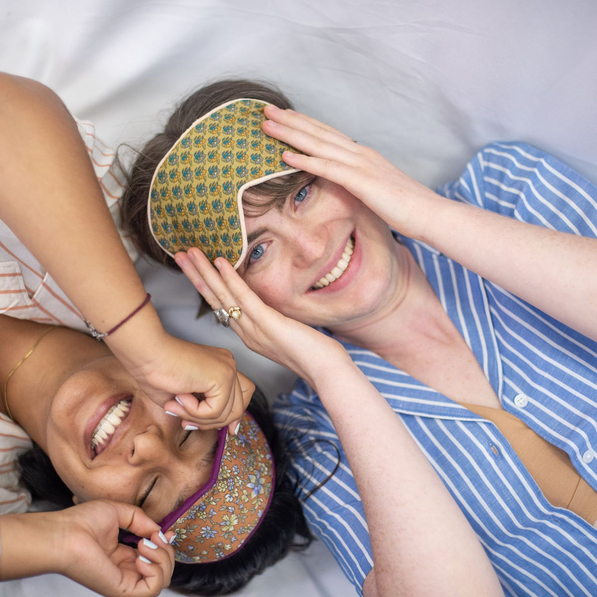 A close up, birds-eye picture of two women wearing the YOU Underwear woven pyjamas and floral eye masks. They are laying down on a white sheet and are looking up at the camera, smiling. The woman we can see most of in the pictures is wearing the blue and white striped pyjamas and the woman off to the left is wearing the soft cream and coral stripes.