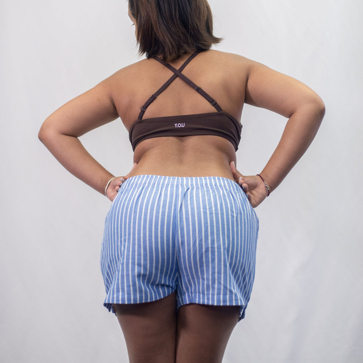 A photo of a model wearing the YOU Underwear woven pyjama shorts in the classic blue and white stripe paired with the cocoa bralette. She is facing away from the camera and leaning forward with her hands on her hips. There is a white background.