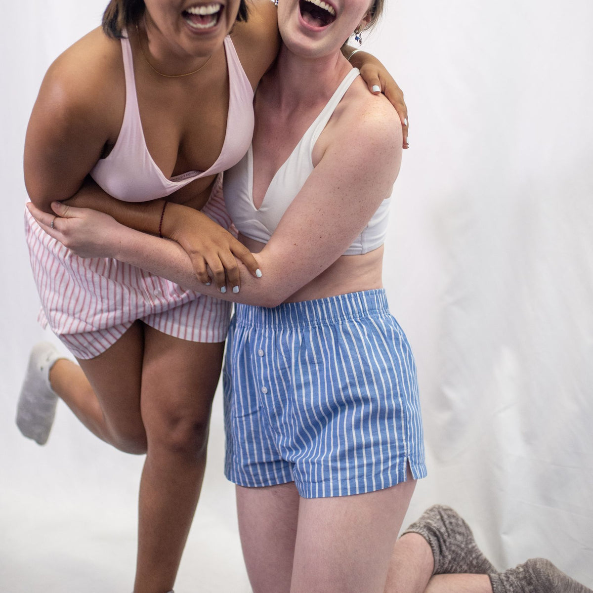 A close up photo of two women wearing the YOU Underwear bralettes and the woven striped pyjama shorts. The model on the right is knelt on the floor and wearing the blue and white striped pyjama shorts. The mode on the left is standing and wearing the pink and white pyjama shorts. They are embraced in a hug and appear to be laughing. There is a white background behind them.