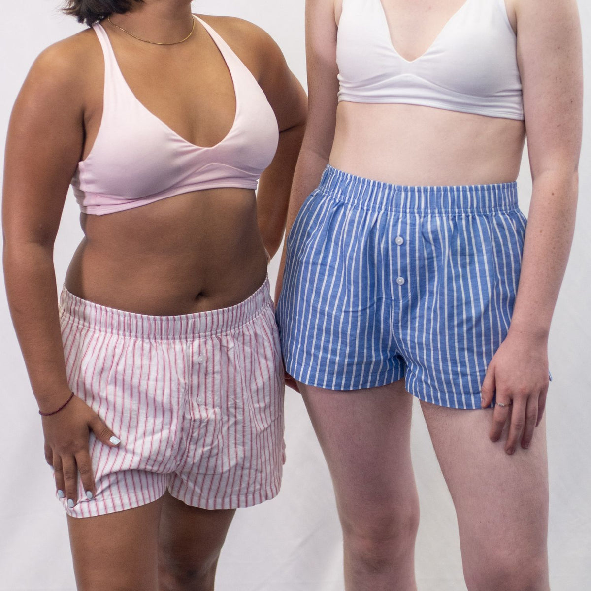 A close up photo of two models who are wearing the YOU Underwear woven pyjama shorts. The woman on the left is wearing a pink bralette and white and pink stripe shorts and the woman on the right is wearing a white bralette and blue and white stripe pyjama shorts. There is a white background behind them and they are both standing facing the camera.
