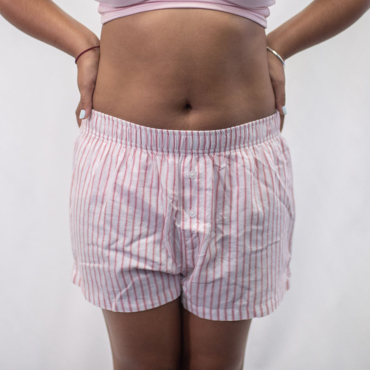A photo of a model wearing the YOU Underwear woven pyjama shorts in white with pink and red stripes. The photo is zoomed in so we can see from the model's stomach to their upper-thigh. She is facing the camera and has her hands on her hips. There is a white background. 