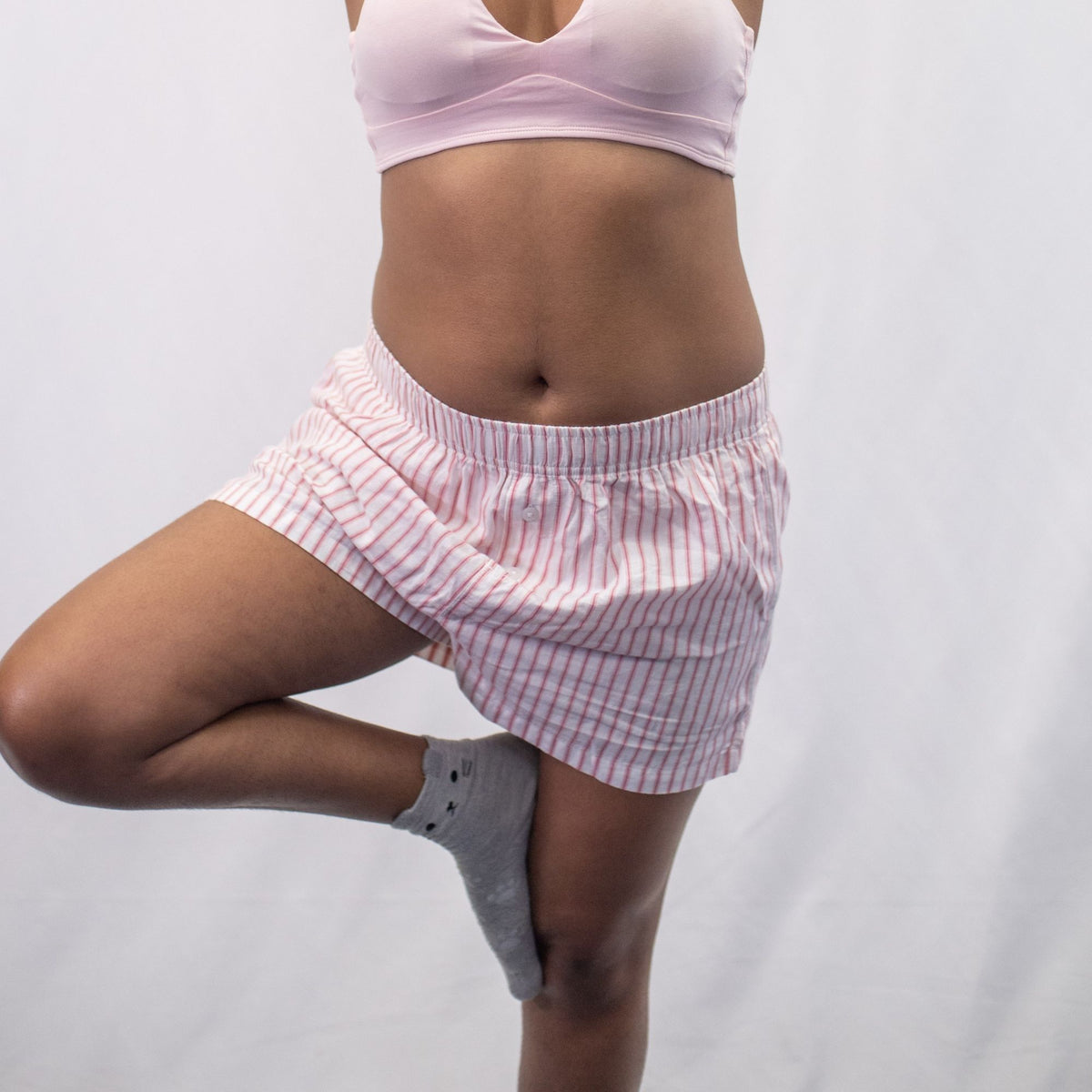 A photo of a model wearing the YOU Underwear woven pyjama shorts in white with pink and red stripes. The photo is zoomed in so we can see from the model's stomach to their upper-thigh. She is standing facing the camera, balancing on leg as if in the tree yoga pose. There is a white background. 