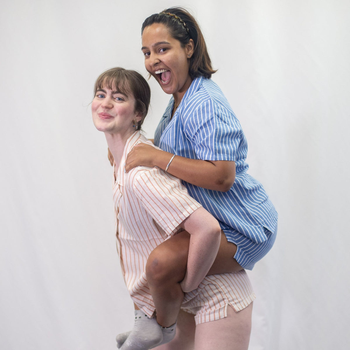 A photo of two models wearing the YOU Underwear woven pyjama shirts and matching shorts. One model, wearing the coral and cream stripe pyjamas, is giving a piggy back to the other model who is wearing the classic blue and white stripe pyjamas. They are smiling and looking at the camera. There is a white background.