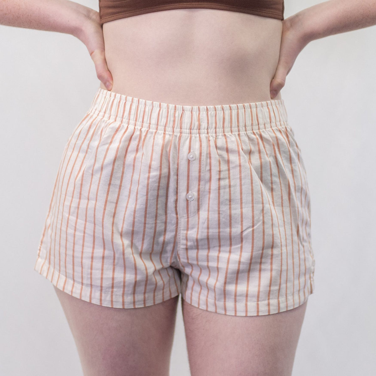 A photo of a model wearing the YOU Underwear woven pyjama shorts in the soft cream and coral stripes. The photo is zoomed in so we can see from the model's stomach to their upper-thigh. She is facing the camera and has her hands on her hips. There is a white background. 