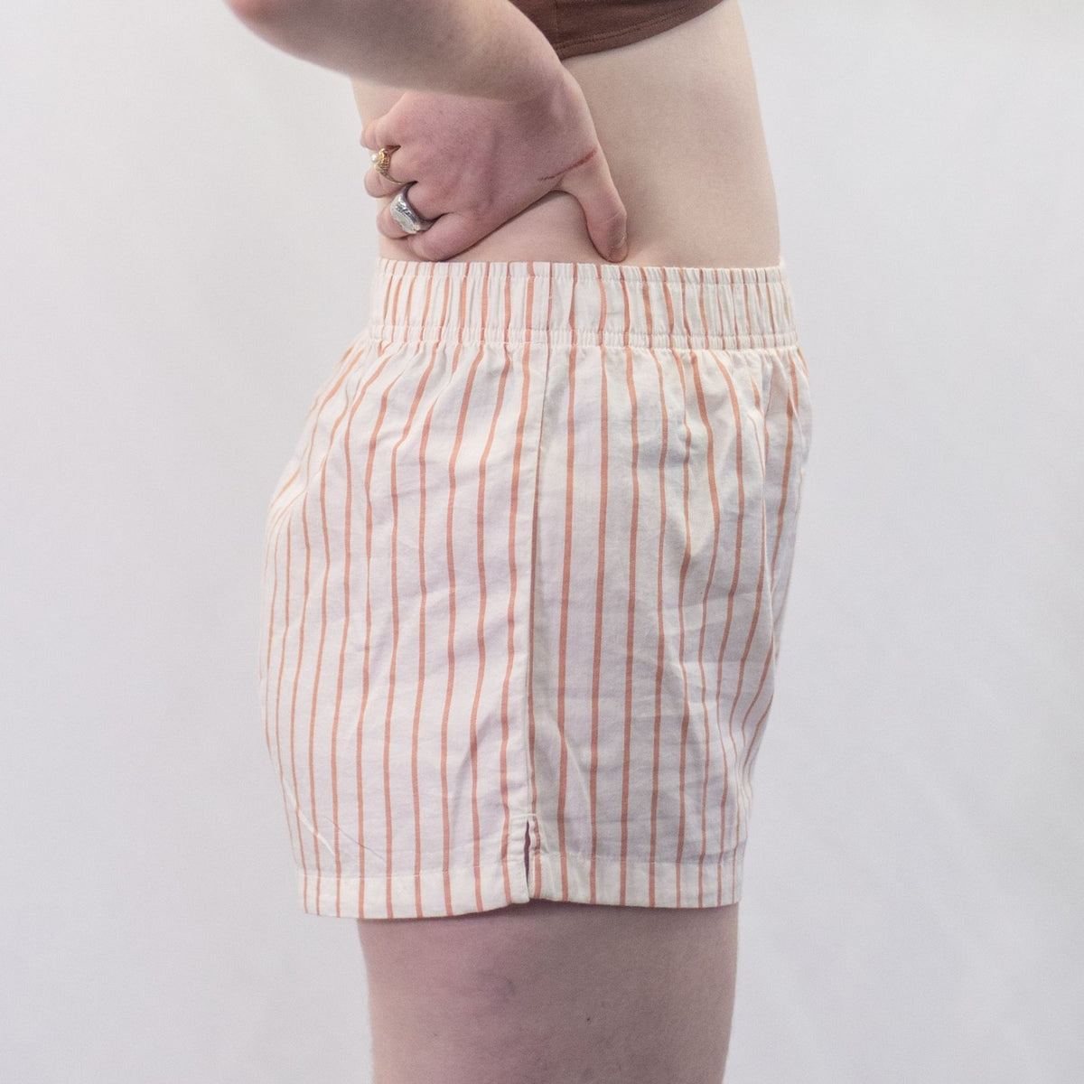 A photo of a model wearing the YOU Underwear woven pyjama shorts in the soft cream and coral stripes. The photo is zoomed in so we can see from the model's stomach to their upper-thigh. She is standing side on to the camera and has her hands on her hips. There is a white background. 