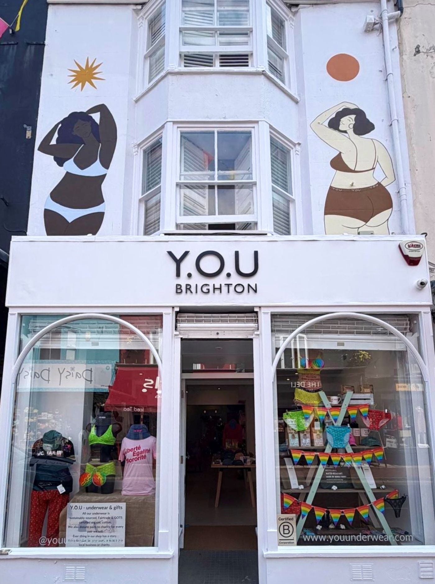 A photo of the YOU Brighton shop front. The building is white with two women's bodies painted on the upper shop front. There is a central door, with two large windows either side of it and a black sign which reads 'YOU Brighton.' The windows are colourful, showcasing the underwear and with pride decorations. 