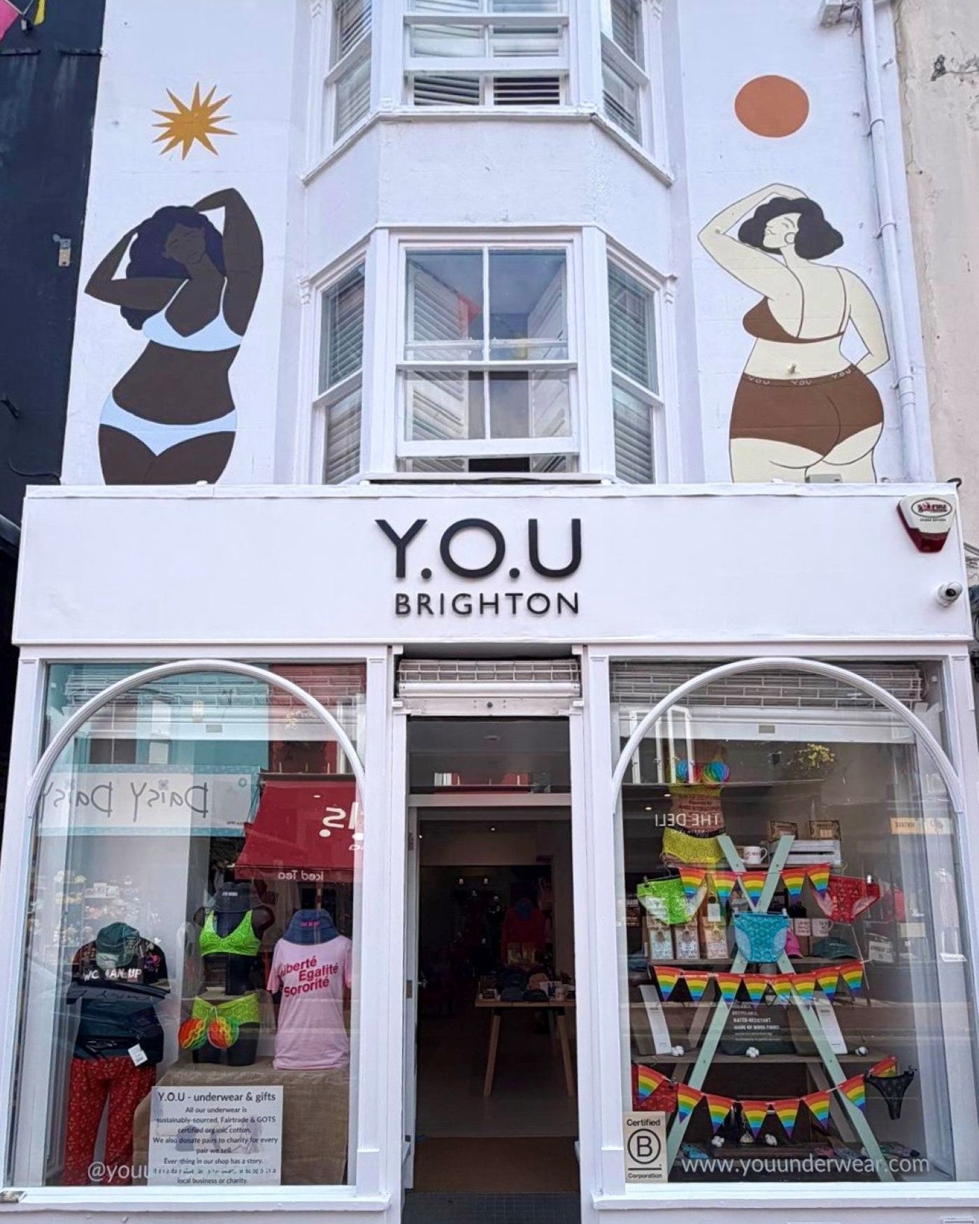 A photo of the YOU Brighton shop front. There are two bodies, wearing underwear which have been painted on the front of the building. There is a central door and two large windows either side which have colourful displays. There is a bold sign which reads, 'YOU Brighton,' on the front of the shop.