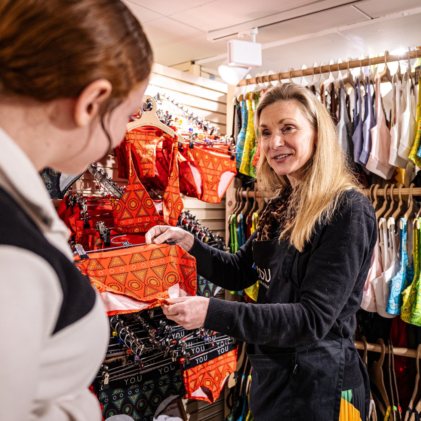 A photo of a YOU Underwear employee showing a customer some red pants. They are smiling and look engaged in conversation. There is lots of colourful underwear in the background, hung on neatly on rails.