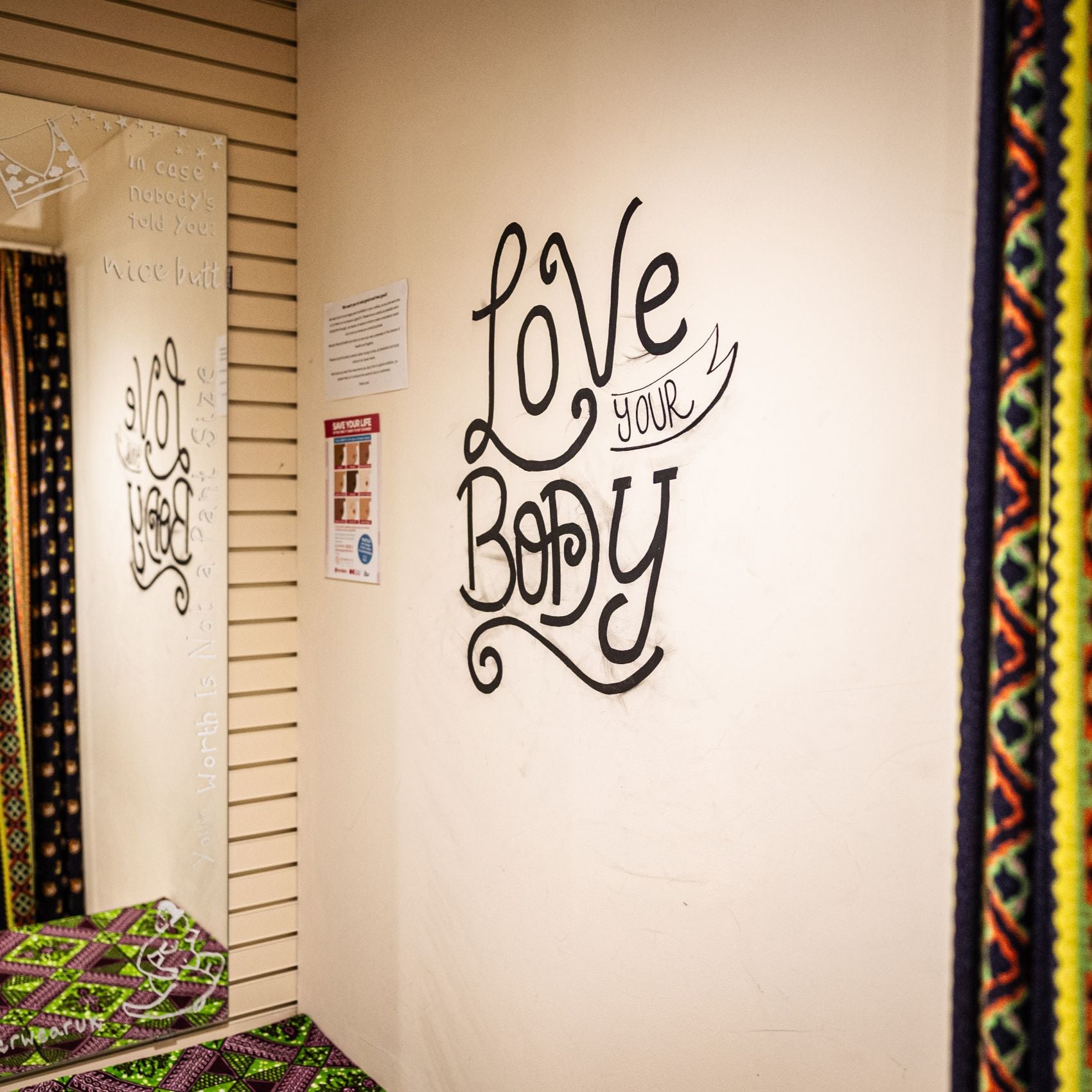 A photo of the inside of one of the YOU Oxford shop's changing rooms. There is a mirror and on the wall there is script style writing which reads, 'Love your body'. The walls are cream and there is a brightly coloured yellow, green and orange curtain in the foreground.