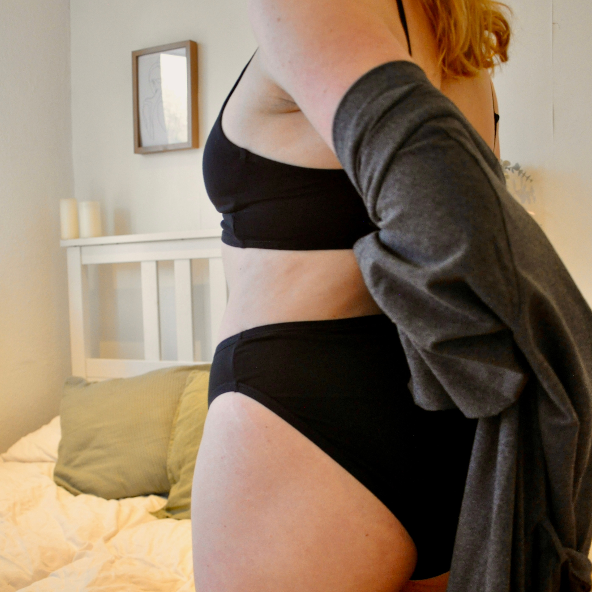 A person standing in a bedroom, wearing black mid-rise bikini underwear and wrapping themselves in a grey robe. The background features a white bed with light bedding and pillows, along with minimalistic wall decor, including a framed picture and two candles.