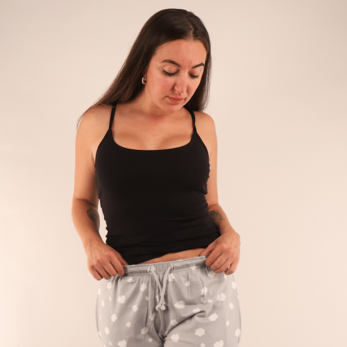 A picture of a woman wearing the YOU Underwear black camisole in black and the grey with clouds pyjama bottoms. She is facing the camera, holding the waistband of her trousers and looking down at them. There is a cream background.