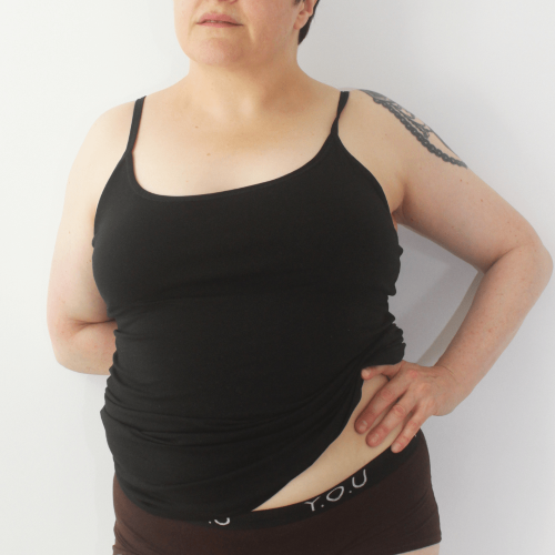 A picture of a model wearing the YOU Underwear black camisole paired with the YOU Underwear branded cocoa boyshorts. The photo is zoomed in so we can see from the model's lips to their hips. They are facing the camera, with their left hand on their hip, slightly pulling up the camisole.