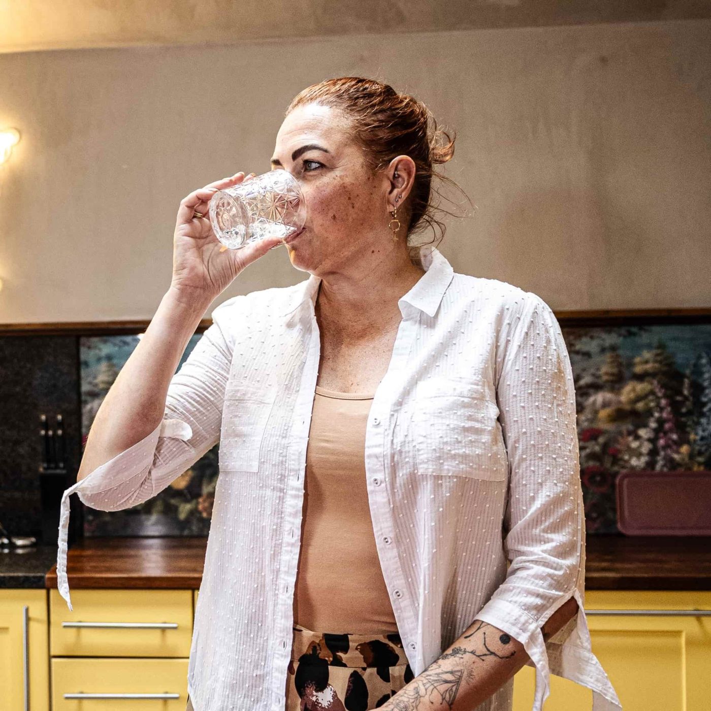 A picture of a woman wearing the YOU Underwear almond vest top paired with an open, button down white shirt and leopard print bottoms. She is sipping from a clear crystal glass, looking out to her right.