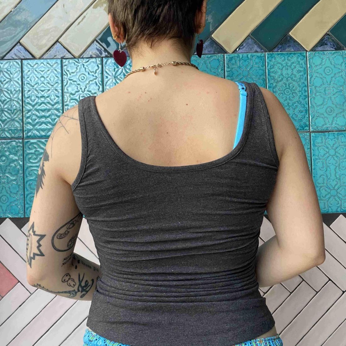 A close up photo of a model wearing the YOU Underwear vest in charcoal grey. They are facing away from the camera. They are stood in front of a white, pink and blue mosaic wall.