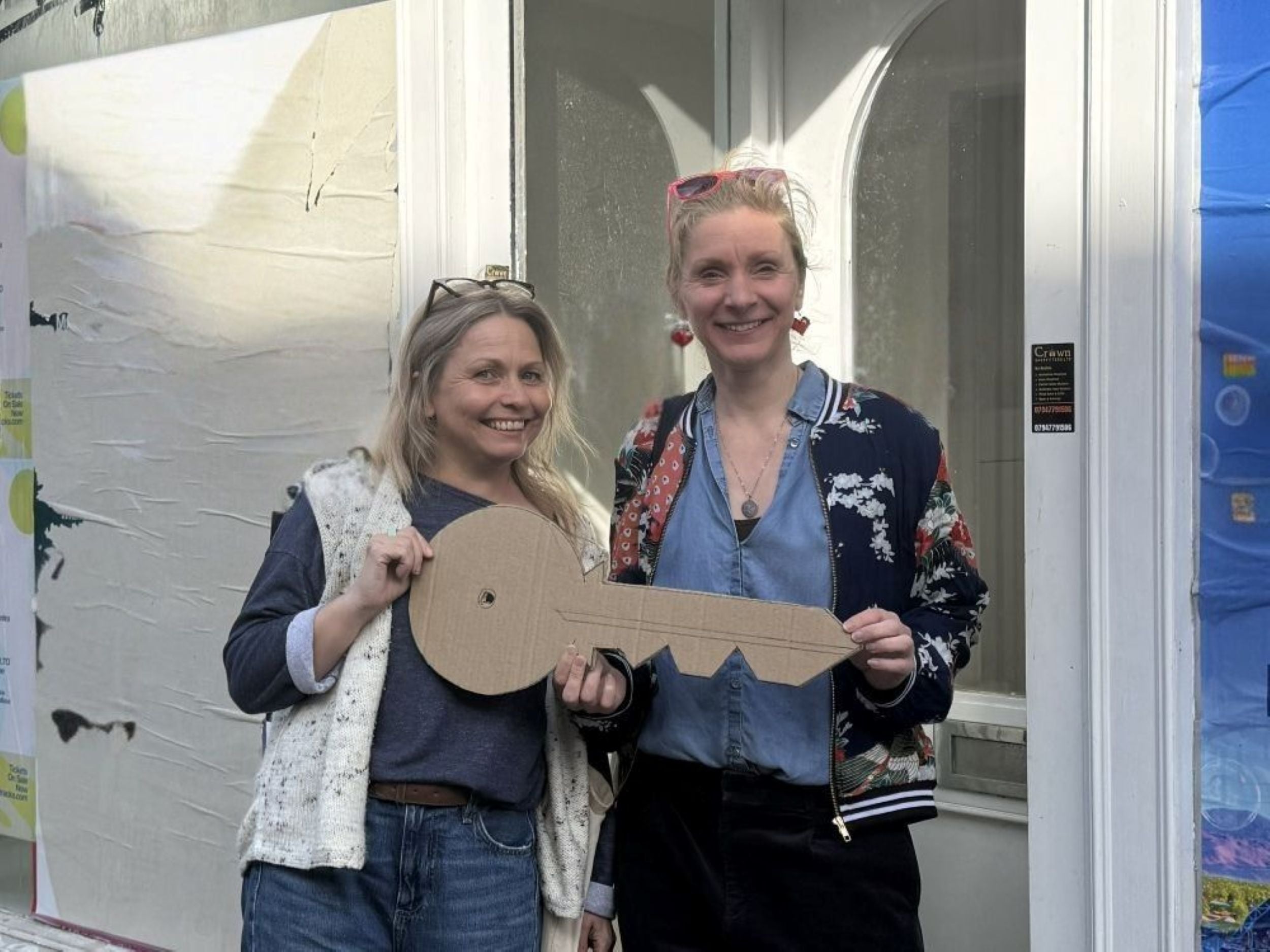 The image shows two women, Denise & Sarah, standing outside an empty shop front holding a large cardboard key