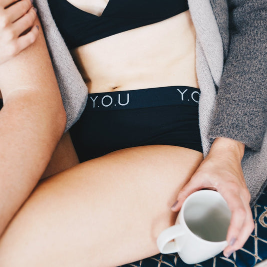 A woman wears black branded boy shorts with a black bralette, she is holding a mug