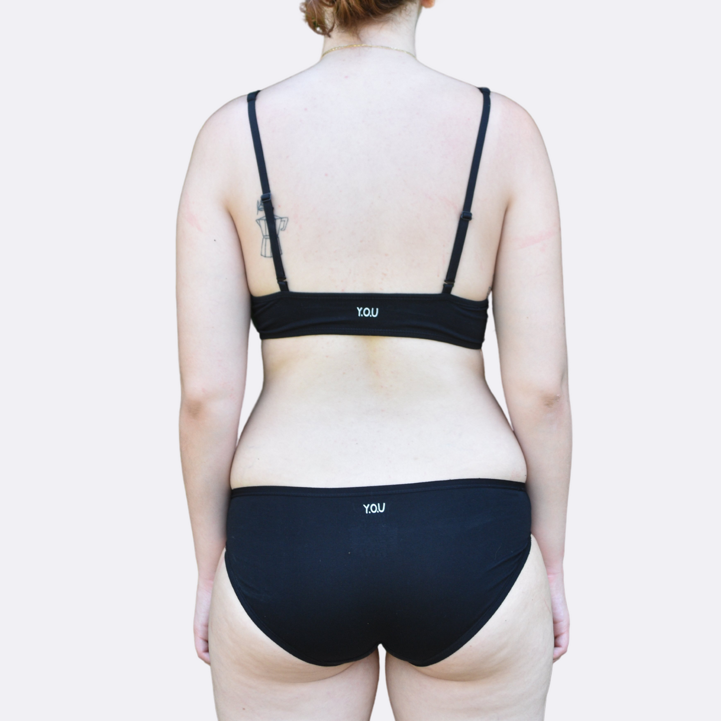 A light skinned woman wears the organic cotton Black bralette and bikini set - facing forward with the back of underwear showing