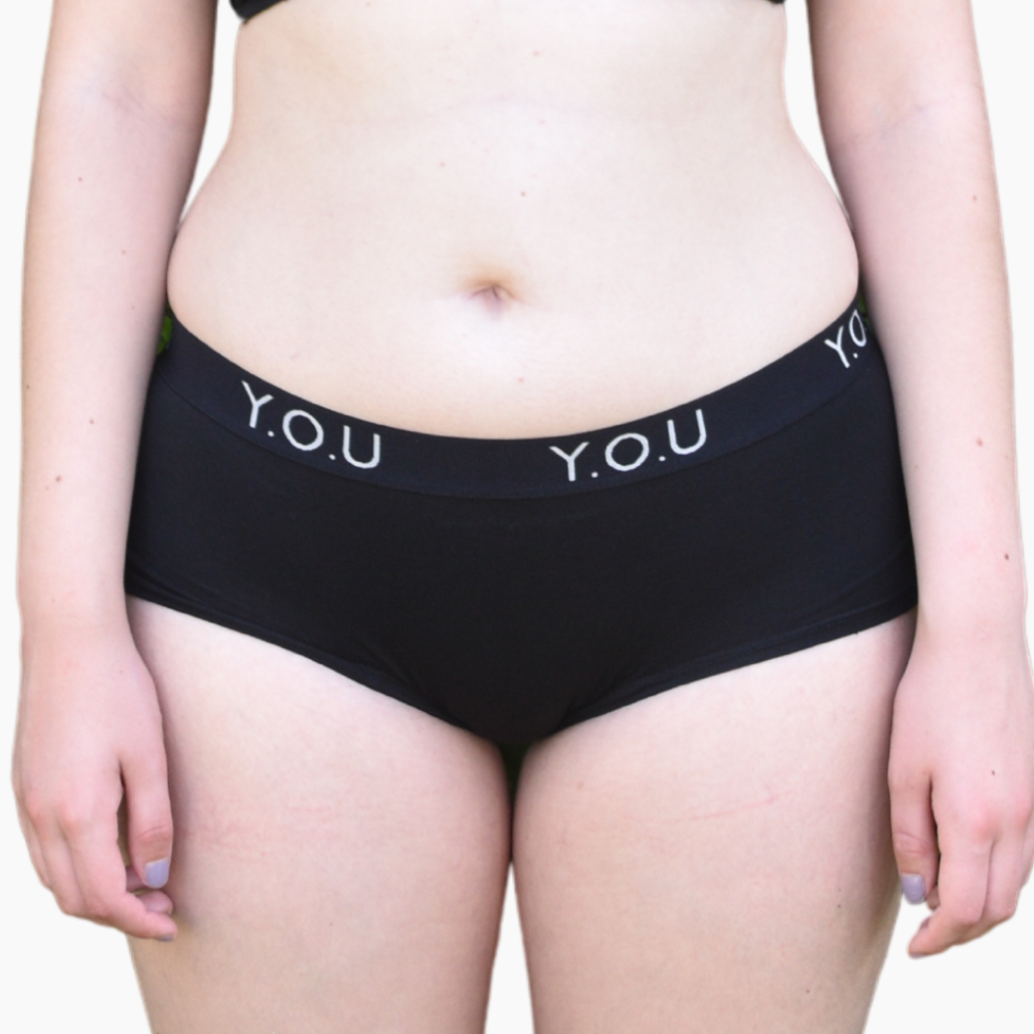 A light skinned woman wears organic cotton black Y.O.U branded boy shorts and stands to the front