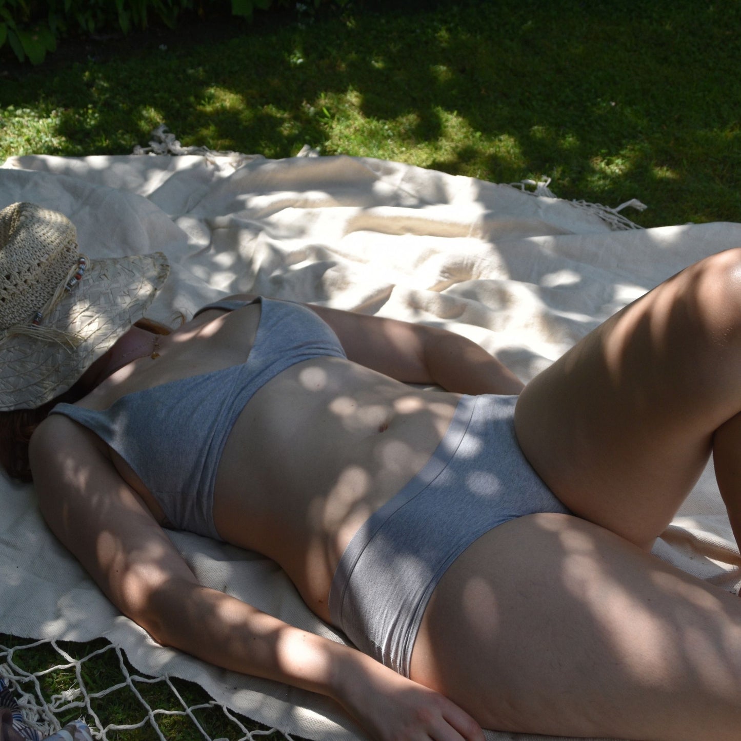  Grey Boy Shorts and Bralette Matching Set Lifestyle  shot - a woman wears the matching set laying on a blanket outside. Her face is covered by a straw hat and the light is dappled on her skin -hover
